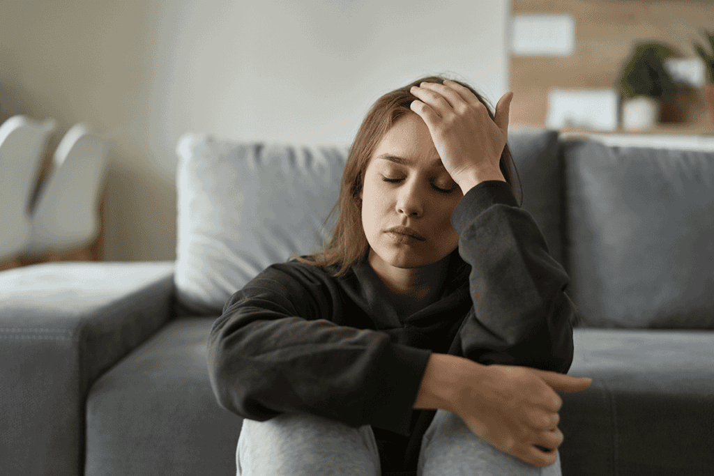 Person sitting on floor against couch with hand on head experiencing signs of alcohol abuse